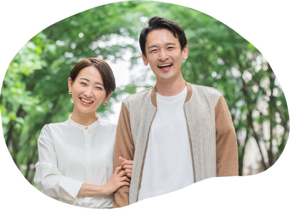緑の木々が見える屋外の公園で、笑顔の若い夫婦二人が寄り添い、正面を見ている様子